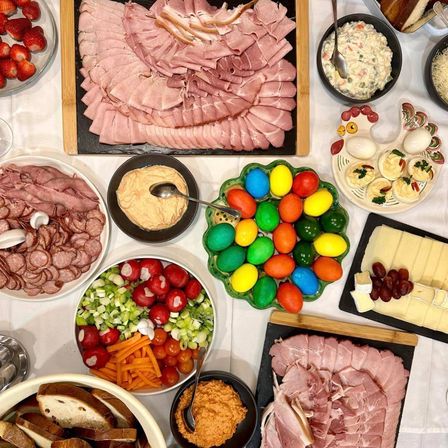 Ein Tisch ist mit verschiedenen Lebensmitteln gefüllt, darunter geschnittener Schinken, bunte Ostereier, eine Dip-Schüssel und eine Platte mit gefüllten Eiern.