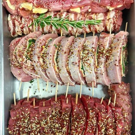 Ein Tablett mit aufgespießten Schweinelenden, jede in Speck und Rosmarin gewickelt, mit verschiedenen Gewürzen bestreut.