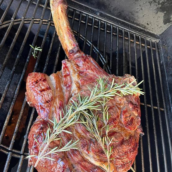 Ein gut gegarter Ribeye-Steak liegt auf einem Grill, garniert mit frischem Rosmarin. Der Steak hat eine reiche, rotbraune Farbe und eine knusprige Außenseite.