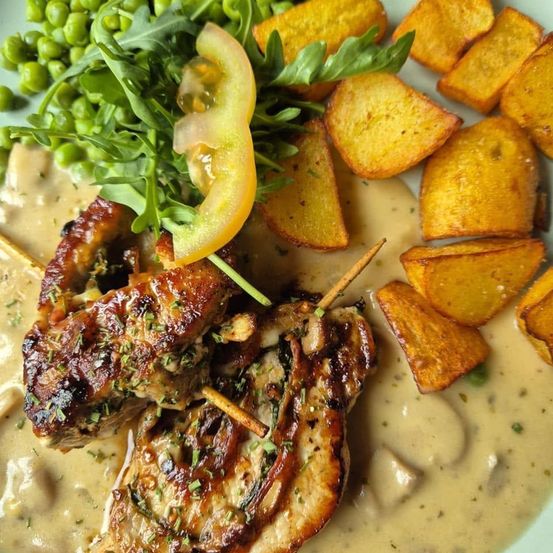 Ein Teller mit gegrillten Hähnchenspießen in cremiger Sauce, gebratenen Kartoffeln und einer Beilage aus grünen Erbsen und Rucola.