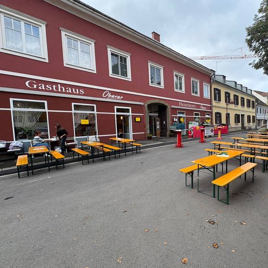 Ein Bereich mit gelben Tischen und Bänken ist vor einem roten Gebäude mit der Aufschrift 'Gasthaus Oberer' eingerichtet. Der Bereich ist derzeit leer, mit einigen Barrieren und einem Kran im Hintergrund.