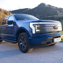 Ein blauer Ford F-150 Pickup-Truck steht auf einer Schotterstraße mit einem Gebirgshintergrund. Die Front zeigt einen kräftigen Kühlergrill und Scheinwerfer, das Kennzeichen SE 996 WS ist sichtbar.