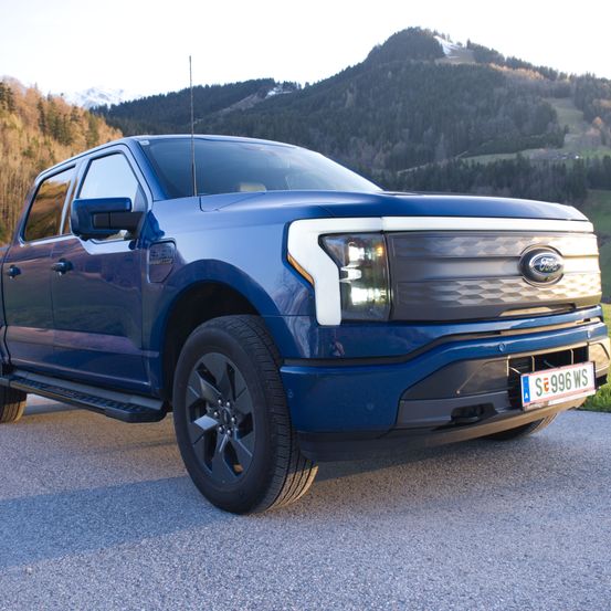 Ein blauer Ford F-150 Pickup-Truck steht auf einer Schotterstraße mit einem Gebirgshintergrund. Die Front zeigt einen kräftigen Kühlergrill und Scheinwerfer, das Kennzeichen SE 996 WS ist sichtbar.