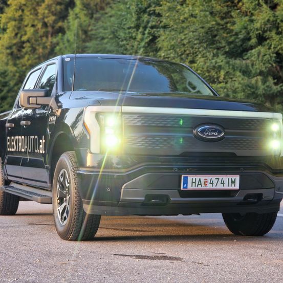 Ein schwarzer Ford F-150 Lightning Elektro-Truck mit eingeschalteten Scheinwerfern ist auf einer Straße mit Bäumen im Hintergrund geparkt.