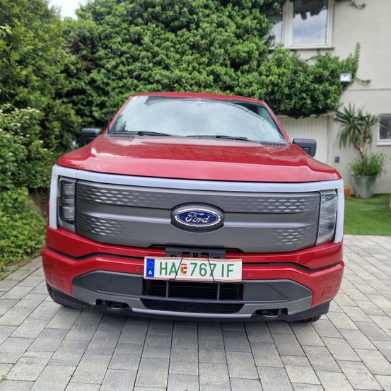Die Vorderansicht eines roten Ford-Fahrzeugs mit einem Nummernschild ist auf einer gepflasterten Einfahrt vor einem Haus mit Grünpflanzen geparkt.