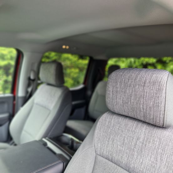 Das Innere eines Fahrzeugs zeigt drei graue Sitze, wobei der Kopfstütze des Vordersitzes im Fokus steht, vor einem verschwommenen Hintergrund von Bäumen.