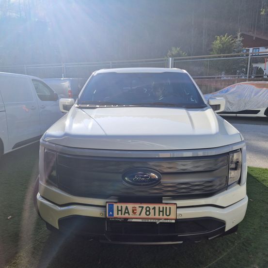 Ein weißer Ford-Fahrzeug ist auf einem Rasenfeld geparkt, mit einem abgedeckten Auto und Lieferwagen dahinter. Das Fahrzeug hat ein Nummernschild mit der Aufschrift HA 781 HU.