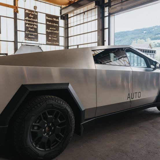 Ein schlankes, metallisches Auto mit dem Wort 'AUTO' an der Seite steht in einer Garage mit Glaswänden. Die Garage ist mit Bannern geschmückt und von einer bergigen Landschaft umgeben.