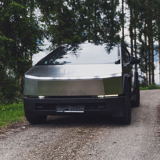 Ein schlankes, futuristisches Auto mit metallischer Oberfläche steht auf einem Schotterweg, umgeben von hohen Bäumen.