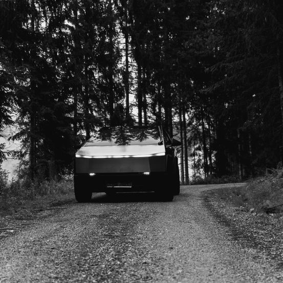 Ein Schwarz-Weiß-Bild zeigt ein schlankes Auto auf einer Kiesstraße, umgeben von einem dichten Wald. Die Scheinwerfer des Autos sind eingeschaltet und beleuchten den Weg vor ihm.
