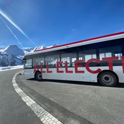 Ein weißer Bus mit roten Buchstaben steht auf einer verschneiten Bergstraße. Im Hintergrund sind Berge zu sehen.