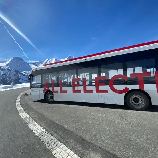 Ein weißer Bus mit roten Buchstaben steht auf einer verschneiten Bergstraße. Im Hintergrund sind Berge zu sehen.