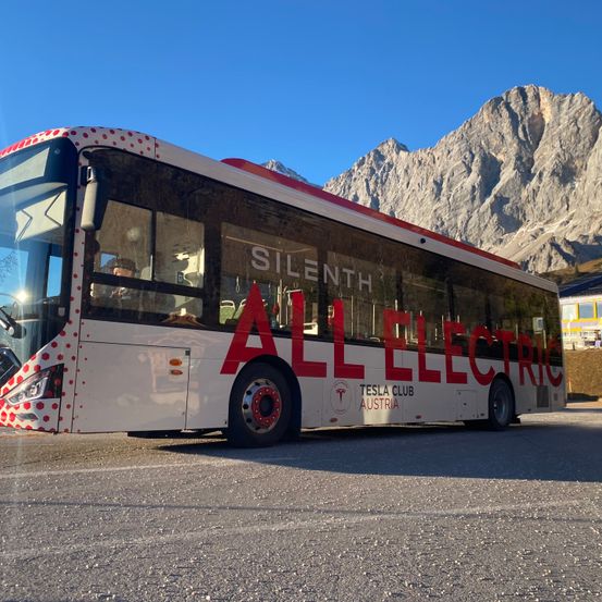 Ein weißer Bus mit roten Punkten und roten Schriftzug 'ALL ELECTRIC' steht vor einer Bergkette.