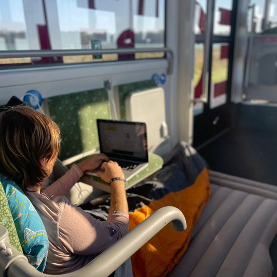 Eine Frau sitzt in einem Stuhl in einem Bus und benutzt einen Laptop auf einer Ablage. Sie hat ein Kissen auf dem Schoß und eine orange Decke neben sich.