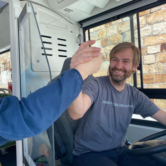 Ein Mann in einem grauen T-Shirt sitzt in einem Bus und hält die Hand einer anderen Person in einer blauen Jacke.