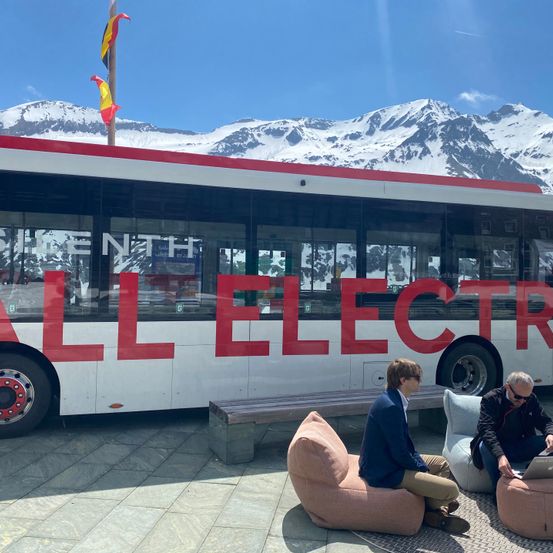 Zwei Männer sitzen auf Sitzsäcken und arbeiten vor einem Bus vor sich auf Laptops. Der Bus steht 'ALL ELECTR'. Eine Flagge und verschneite Berge sind im Hintergrund.