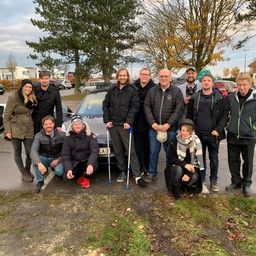 Eine Gruppe von Menschen steht vor einem geparkten Auto und lächelt für ein Foto. Einige benutzen Gehhilfen, und im Hintergrund sind Bäume und geparkte Fahrzeuge zu sehen.