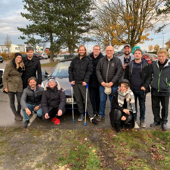 Eine Gruppe von Menschen steht vor einem geparkten Auto und lächelt für ein Foto. Einige benutzen Gehhilfen, und im Hintergrund sind Bäume und geparkte Fahrzeuge zu sehen.