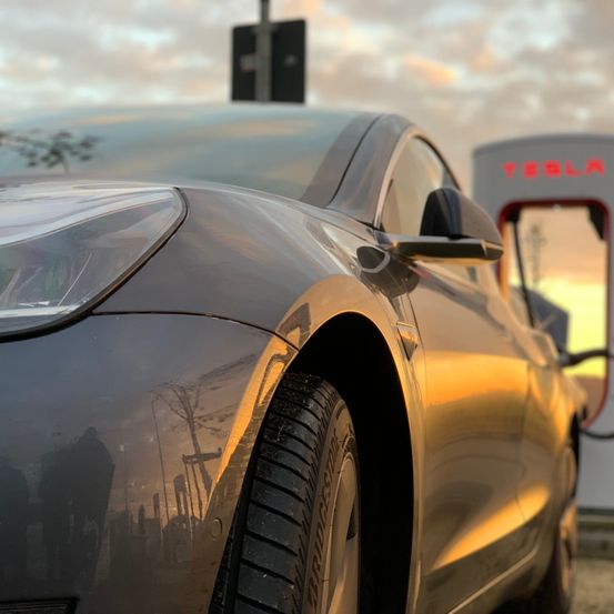 Ein schlanker schwarzer Tesla Model S ist an einer Tesla-Ladestation mit einem Sonnenuntergang im Hintergrund geparkt.