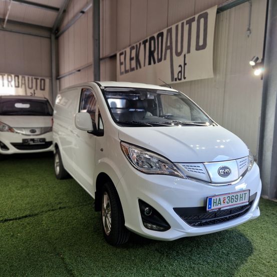 Ein weißer Elektrovan mit dem Kennzeichen HA E 369 HT steht in einer Ausstellungshalle mit grünem Teppich. Ein Banner mit der Aufschrift Elektroauto.lat hängt darüber.