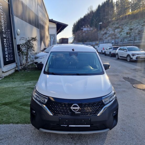 Ein weißer Nissan Elektrovan steht auf der Straße, mit weiteren Fahrzeugen, einem Gebäude und einem Zaun im Hintergrund.