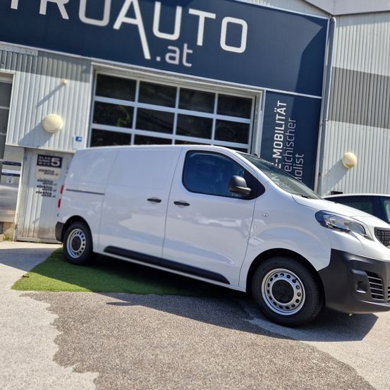 Ein weißer Lieferwagen steht auf grünem Rasen vor einem Gebäude mit der Aufschrift 'RUAUTO.at'. Hinter dem Lieferwagen befindet sich eine große Glastür und eine Wand mit zusätzlichem Text.