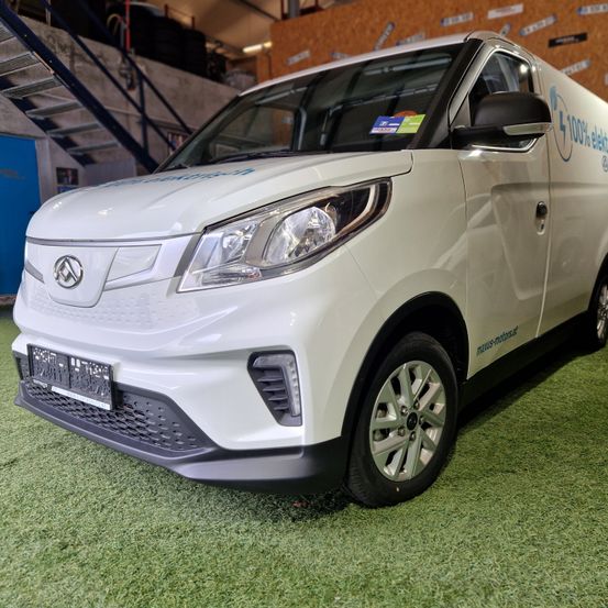 Ein weißer Elektrofahrzeug ist auf einem grünen Teppich geparkt und zeigt seinen vorderen Stoßfänger und Scheinwerfer. Das Fahrzeug hat ein Nummernschild und ein Ladesymbol an der Seite.