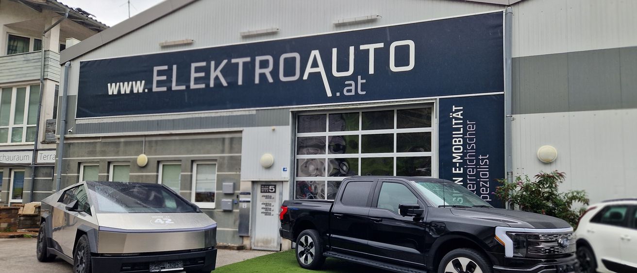 Zwei Autos, ein silbernes und ein schwarzes, parken vor einem Gebäude mit dem Schild Elektroauto.at. Das schwarze Auto ist ein Pickup-Truck.