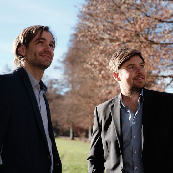 Zwei Männer stehen draußen in einem Park. Beide tragen Anzüge und haben Bärte. Der Mann auf der linken Seite lächelt, während der Mann auf der rechten Seite lächelt und ihn ansieht. Bäume mit roten Blättern sind im Hintergrund.