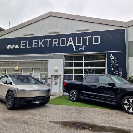 Zwei Elektrofahrzeuge sind vor einem Gebäude mit einem Schild, das 'www.ElektroAuto.at' zeigt, geparkt. Das Gebäude hat auch einen verglasten Eingang mit einer weißen Tür.