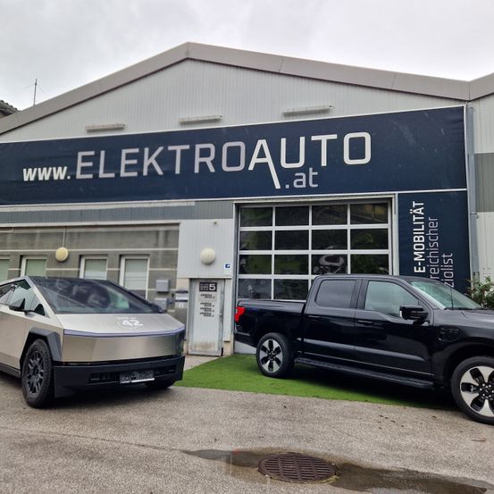 Zwei Elektrofahrzeuge sind vor einem Gebäude mit einem Schild, das 'www.ElektroAuto.at' zeigt, geparkt. Das Gebäude hat auch einen verglasten Eingang mit einer weißen Tür.
