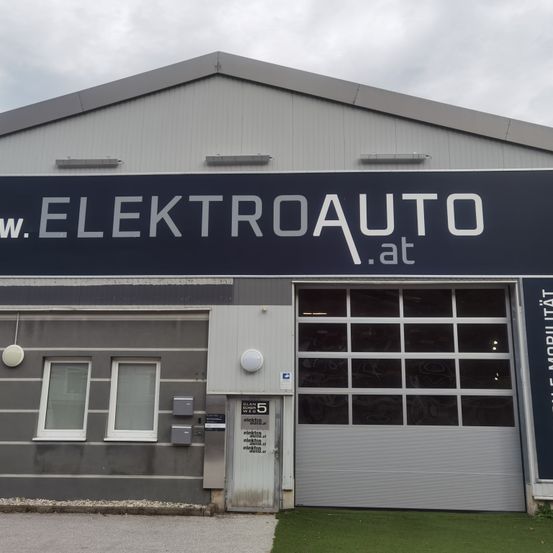 Die Vorderseite eines Industriegebäudes mit der Aufschrift 'w. ELEKTROAUTO .at' mit zwei Fenstern, einer Tür und einer Garagentür unter einem bewölkten Himmel.
