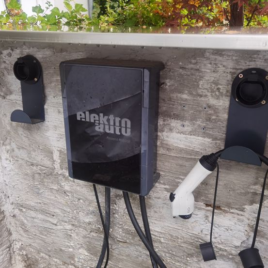 Eine Außenladestation für Elektrofahrzeuge ist an einer Betonwand montiert. Sie hat ein schlankes schwarzes Design mit der Markenbezeichnung 'elektro auto' und einem angeschlossenen weißen Ladekabel.