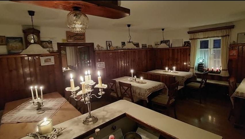 Ein gemütliches Esszimmer mit Holzwänden und -decke. Zwei Tische sind mit brennenden Kerzen besetzt. Es gibt einen Kronleuchter, eine Uhr und Bilderrahmen an der Wand.