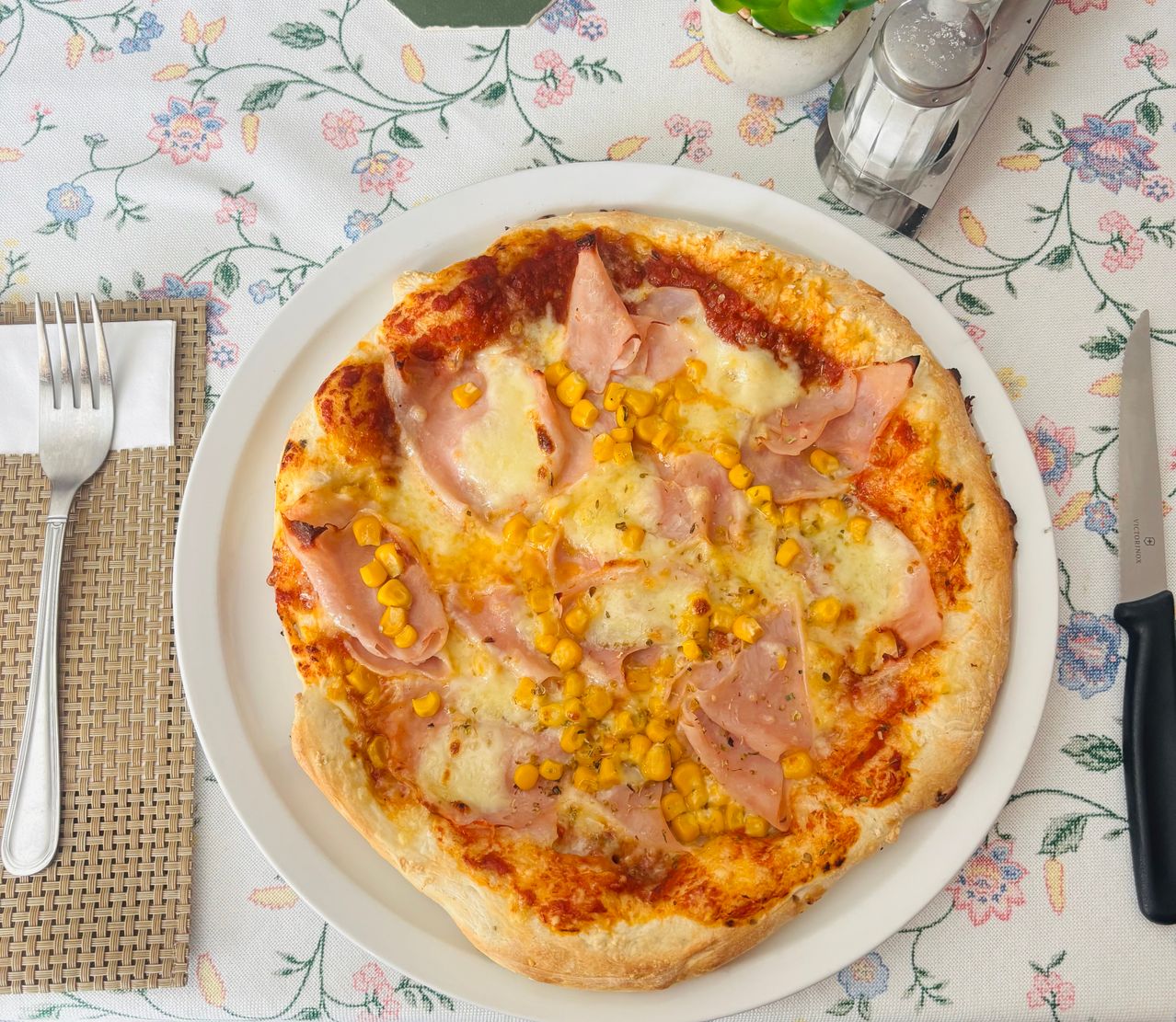 A pizza with ham and corn on a white plate on a floral tablecloth, with a fork and knife on the side.