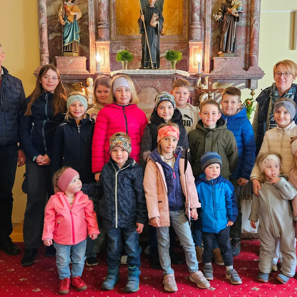 Eine Gruppe von Kindern und Erwachsenen in Winterkleidung posiert vor einem Kirchenaltar mit Statuen, Kerzen und einem roten Teppich.