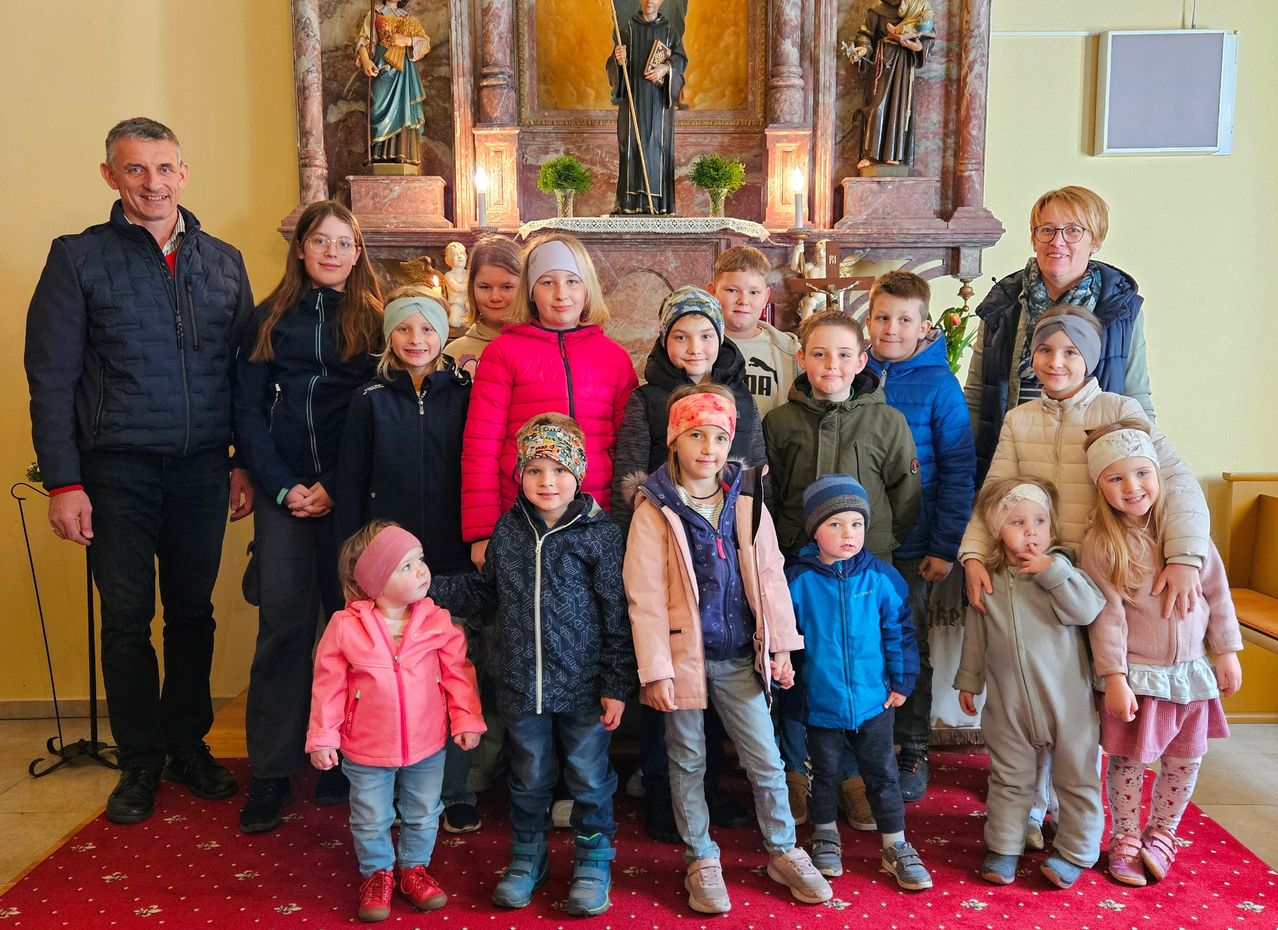 Eine Gruppe von Kindern und Erwachsenen in Winterkleidung posiert vor einem Kirchenaltar mit Statuen, Kerzen und einem roten Teppich.
