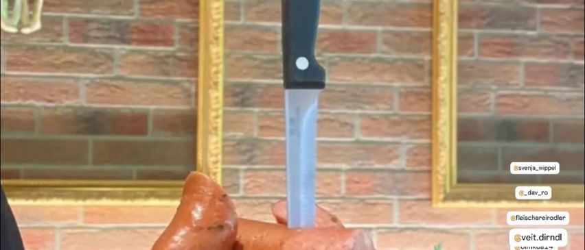 Eine Person hält ein Holzschneidebrett mit Würsten, einem Messer und einer Garnitur darauf. Eine Ziegelmauer und ein goldener Rahmen sind hinter der Person.