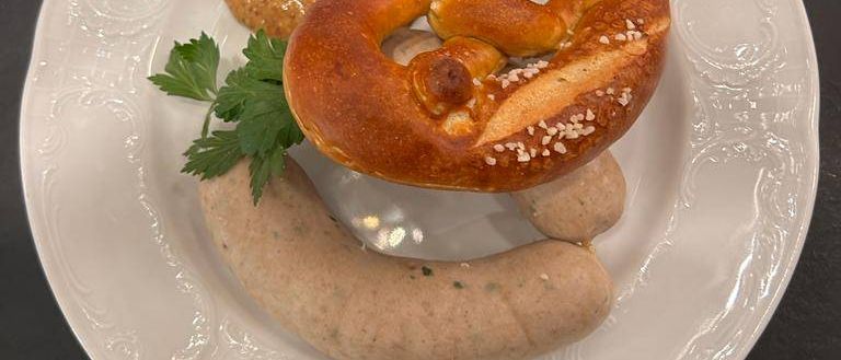 Ein weißer Teller enthält ein Brezel, eine Wurst und eine Dip. Die Brezel hat eine knusprige Kruste und ist mit Sesam bestreut. Die Wurst ist in einer Schüssel.