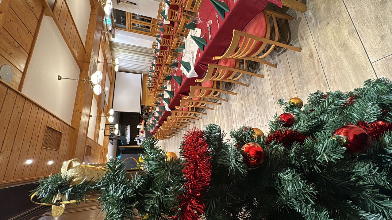 Ein Speisesaal ist mit roten Tischdecken und Servietten, Holzstühlen und einem Weihnachtsbaum im Vordergrund für Weihnachten geschmückt.