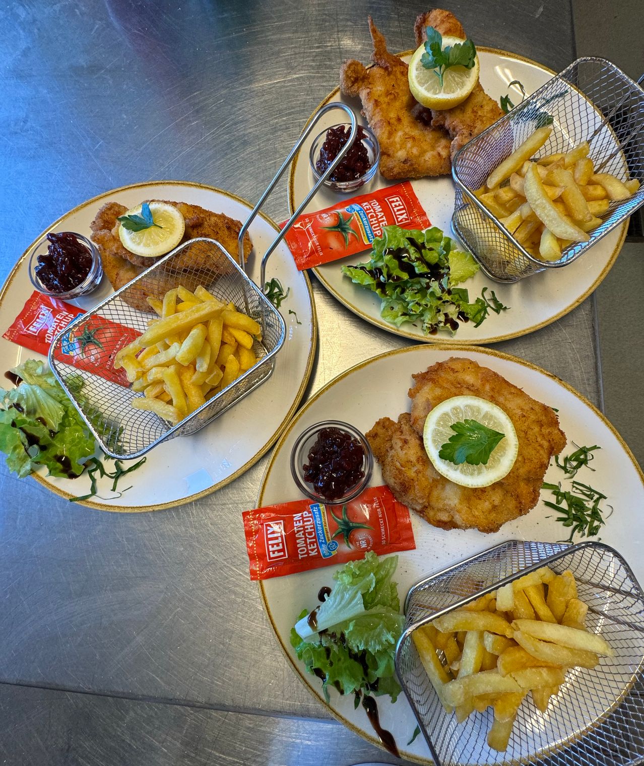 Four plates with fried fish, lemon, and parsley. Each plate has a side of French fries and salad. Condiments like ketchup and jam are also present.