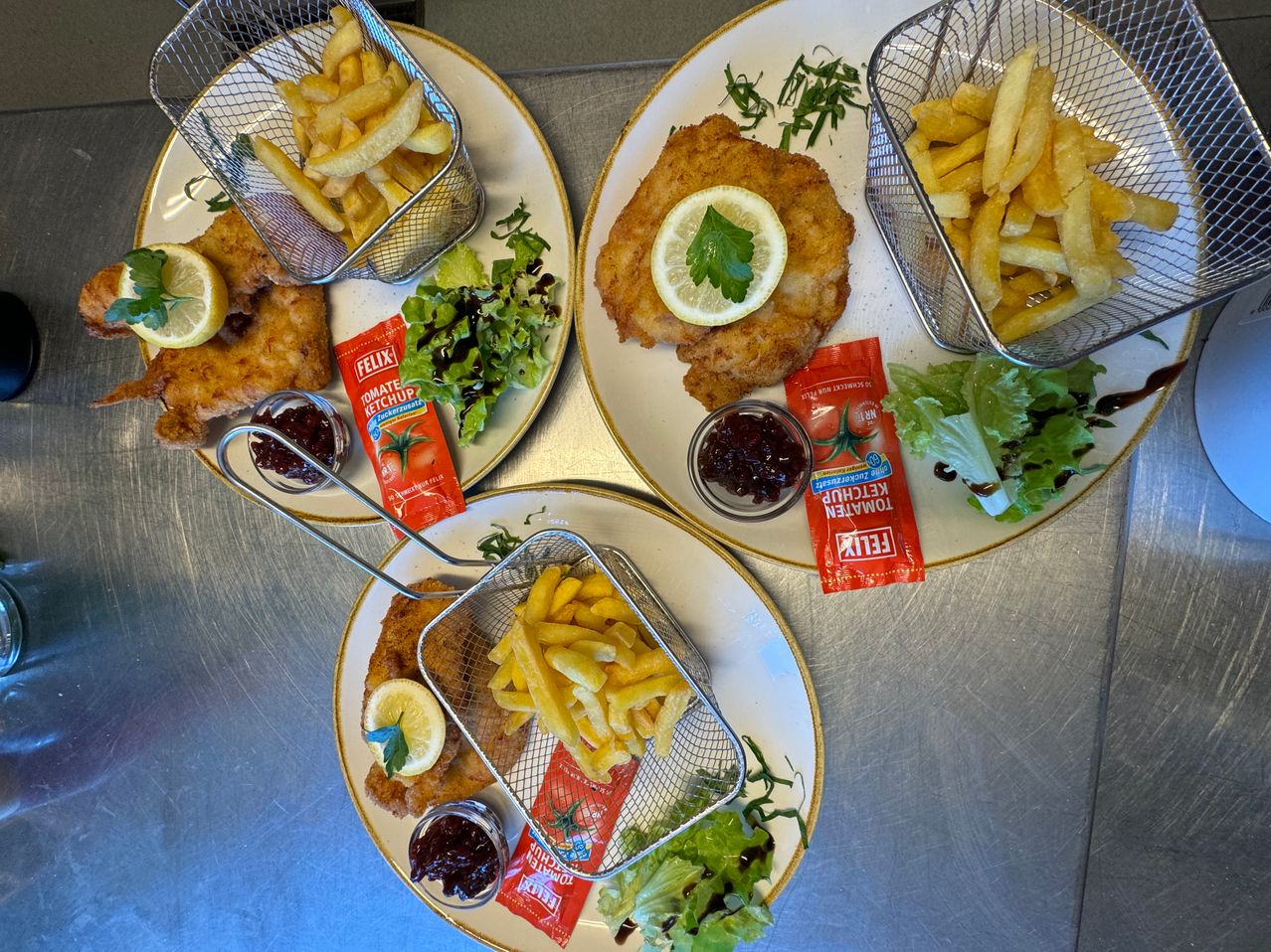 Three plates of food on a table. Each plate contains a fried fish cutlet with a lemon slice and green garnish, accompanied by a basket of French fries and a small container of ketchup.