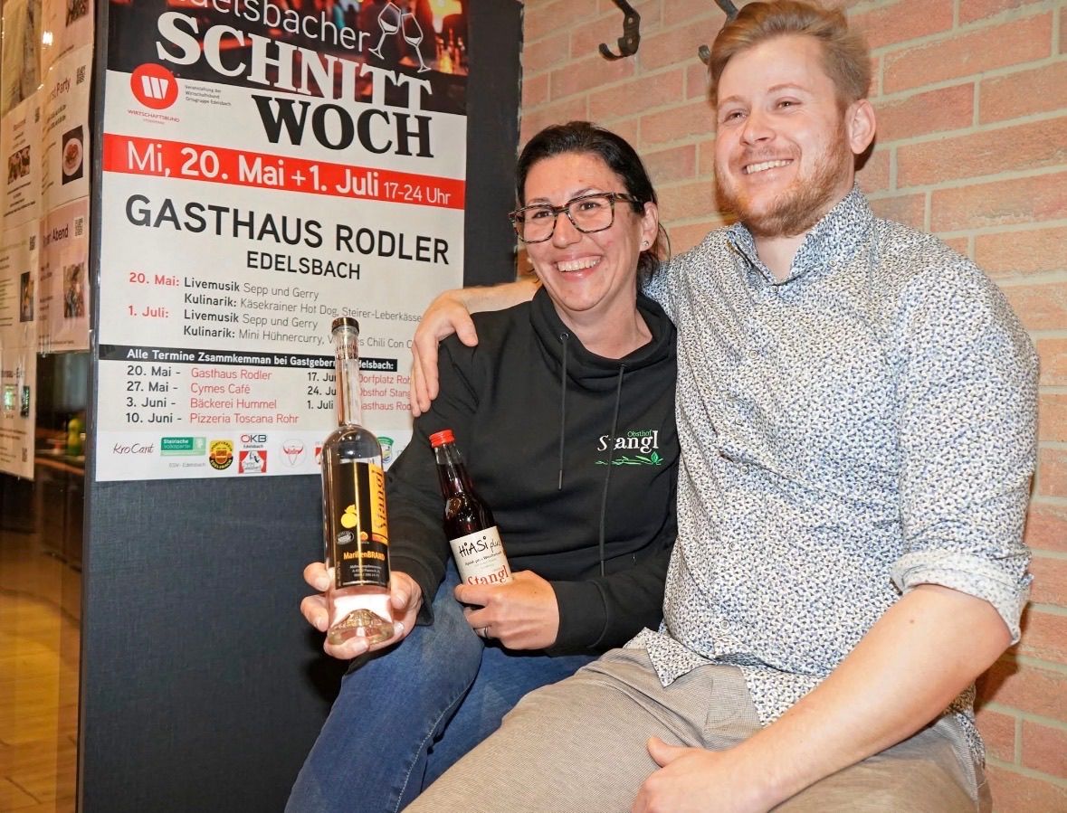 Two individuals seated in front of a brick wall, smiling and holding bottles of spirits. A sign behind them reads Gasthaus Rodler with dates and events listed.