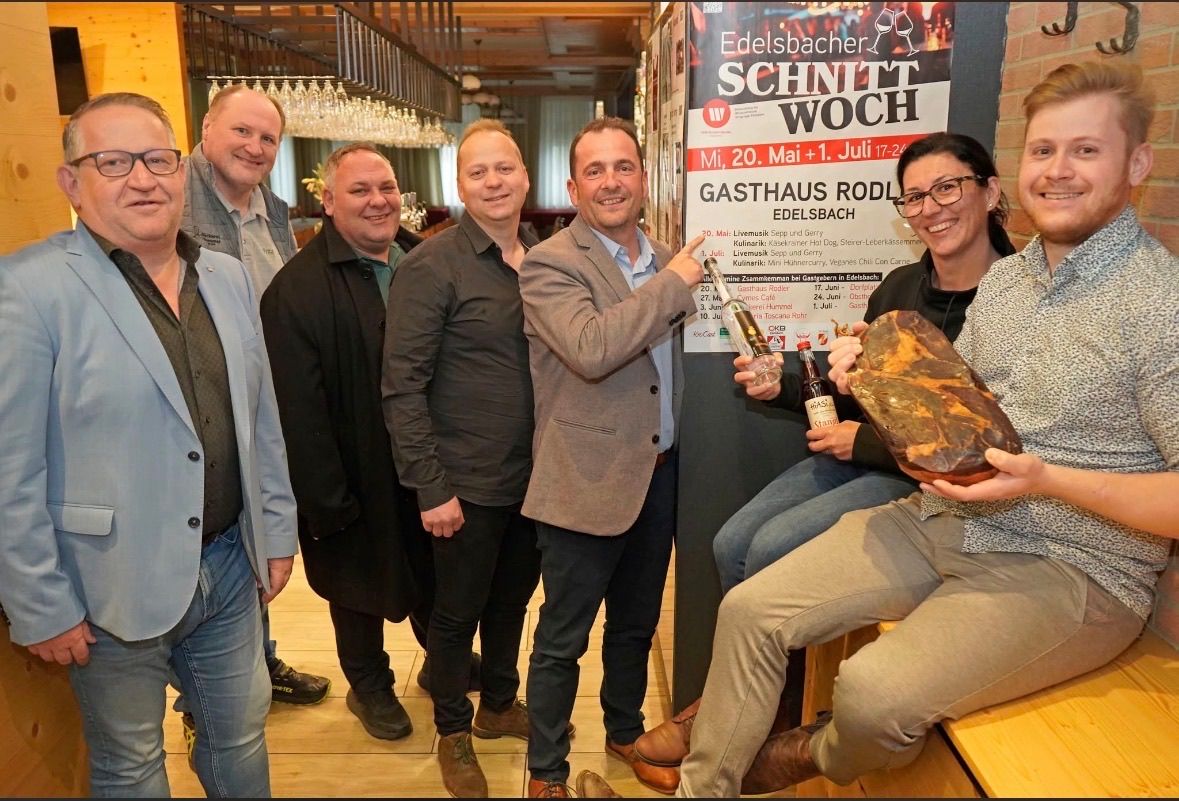 A group of men and women pose for a photo at a restaurant, holding drinks and a large piece of meat. The restaurant has a poster for the 'Edelbacher Schnitt Woche'.