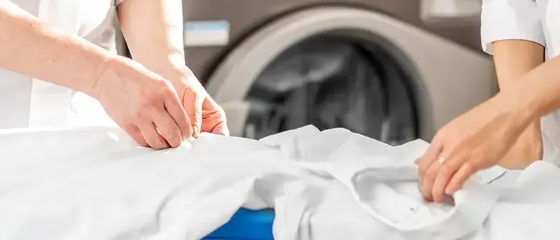Zwei Personen in weißen Uniformen falten ein weißes Hemd auf einem Bügelbrett. Hinter ihnen ist teilweise eine Waschmaschine zu sehen.