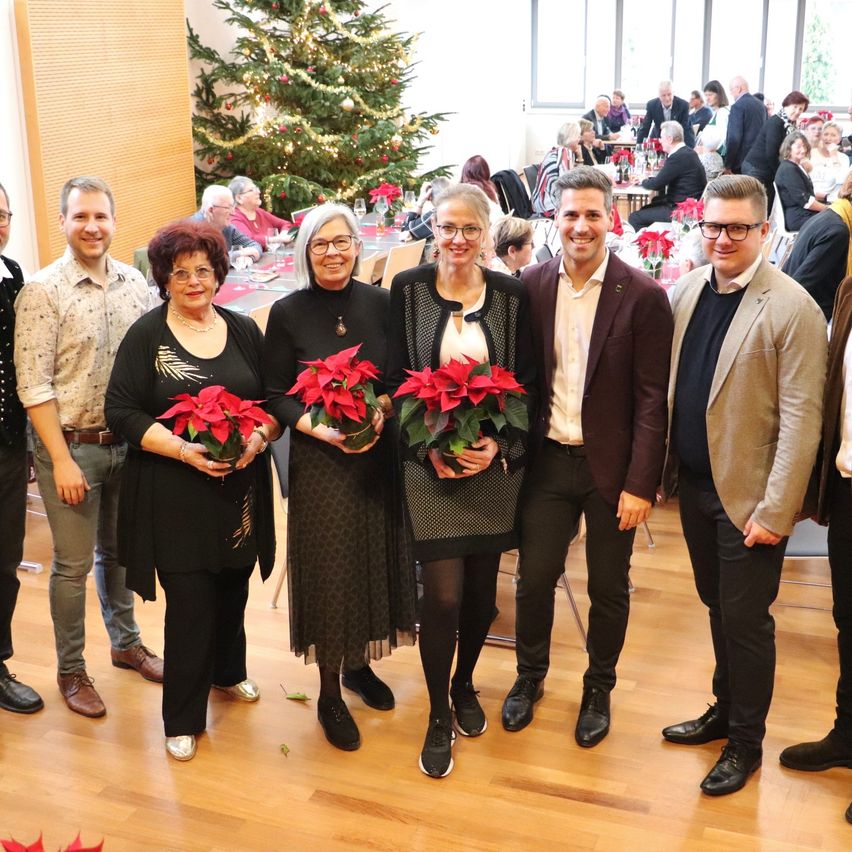 Eine Gruppe von Menschen posiert mit Weihnachtssternpflanzen vor einem geschmückten Weihnachtsbaum in einem Raum.