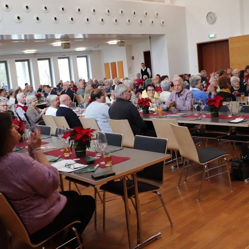 Eine große Versammlung älterer Menschen, die an langen Tischen in einem Speisesaal sitzen, geschmückt mit festlichen Dekorationen und roten Blumen.