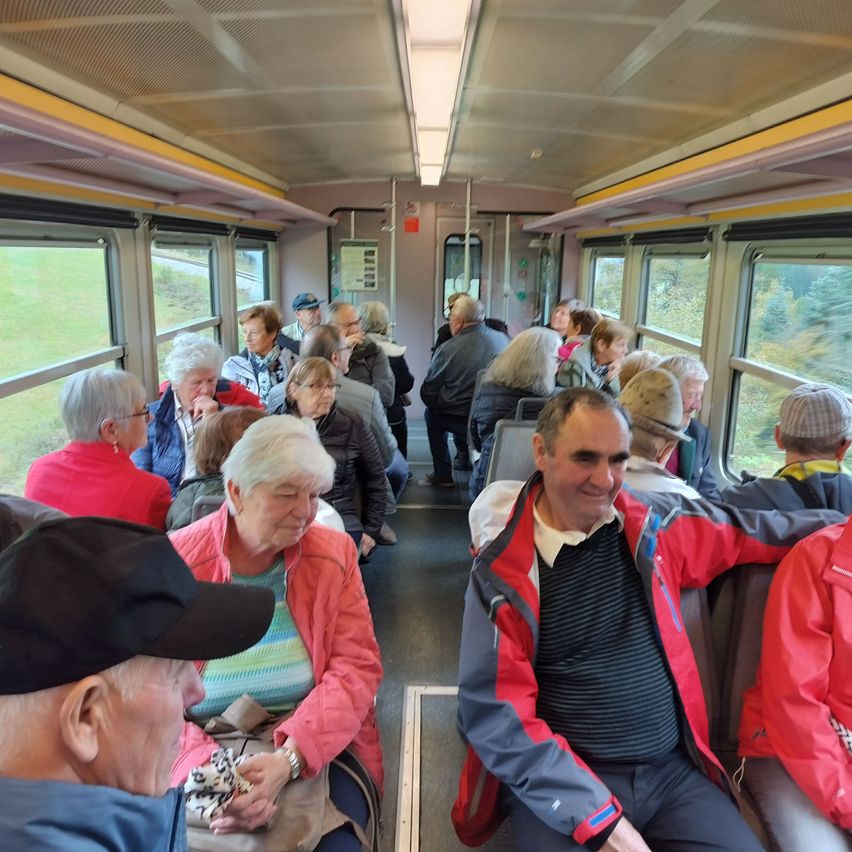 Im Inneren eines Zuges sitzen mehrere ältere Passagiere in Reihen, einige tragen Hüte und Brillen, lächeln und unterhalten sich. Der Zug hat Glasfenster, die ein grünes Feld draußen zeigen.