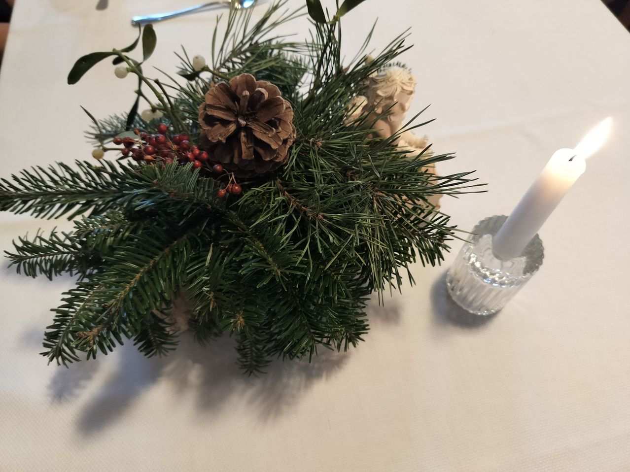 A table setting with a green pine cone centerpiece, surrounded by holly leaves, berries, and a small angel figurine. A candle is next to the centerpiece.