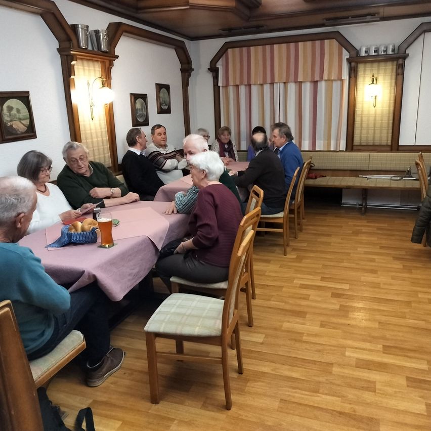 A group of people are sitting around tables in a room. Some are drinking, while others are talking. There are picture frames on the wall and lamps. The floor is wooden.